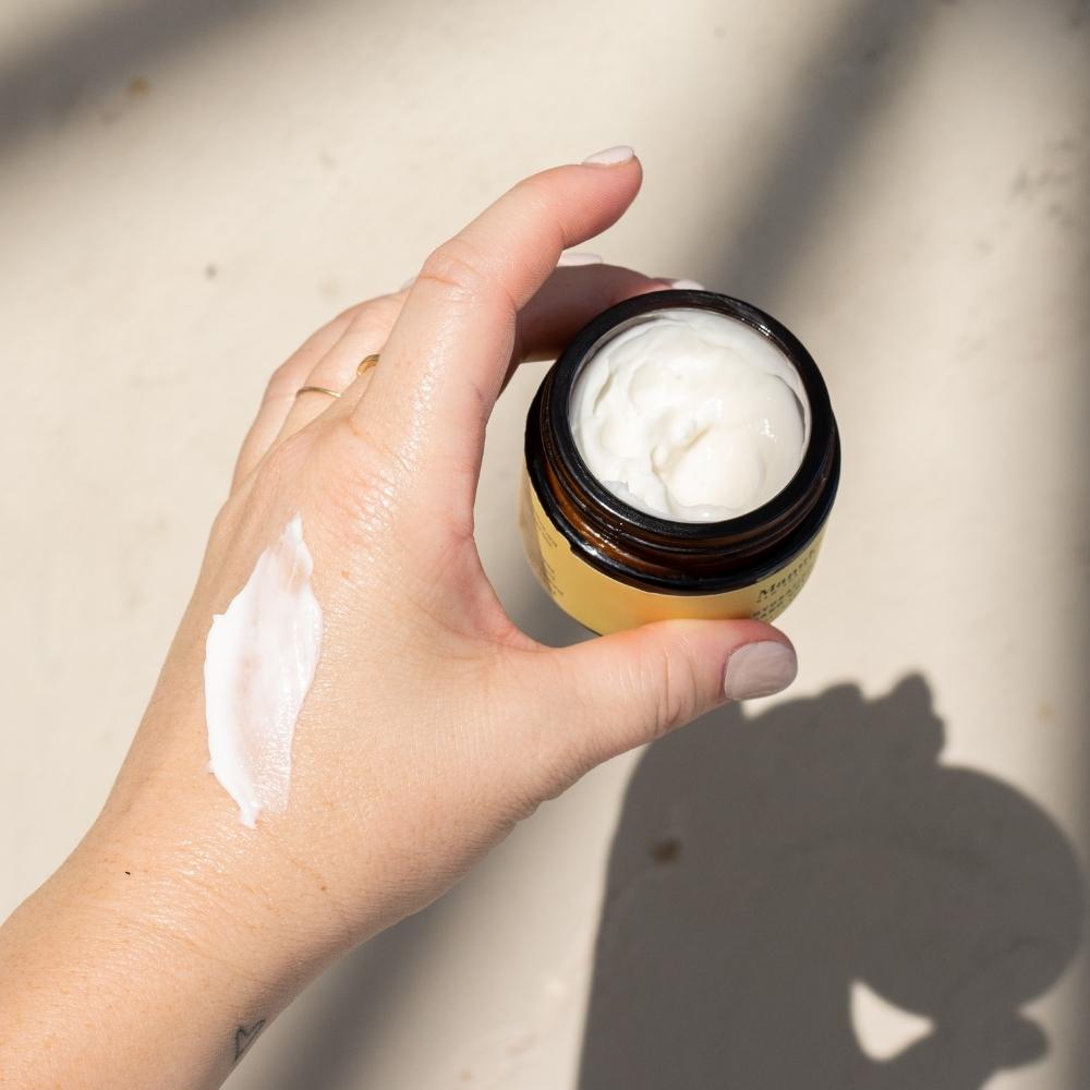 Hand holding open jar of ManukaRx Hydrating Hand Cream with product swatched onto woman's hand